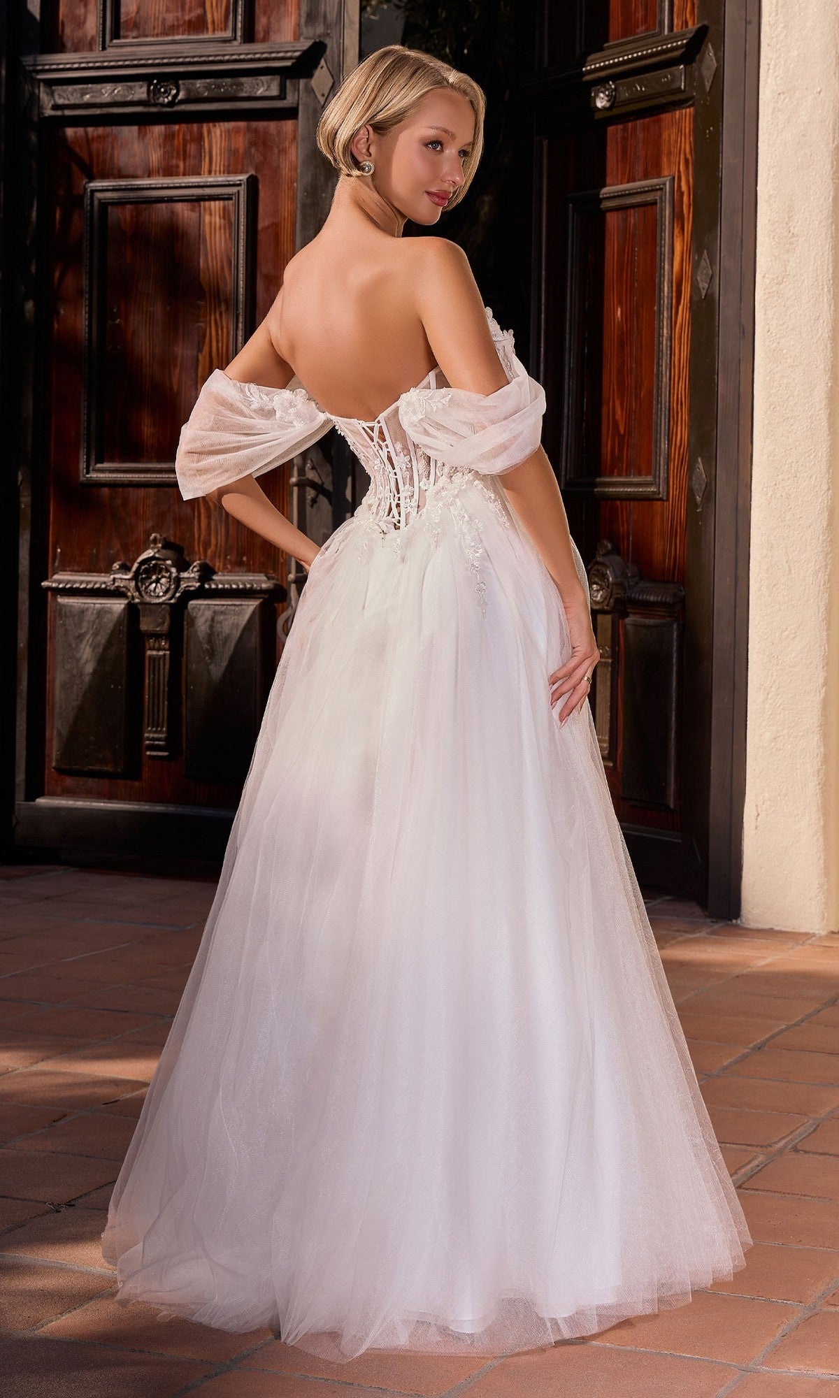 A woman wears the Ladivine CD2426W long formal dress, featuring a white off-the-shoulder illusion corset bodice, posing outdoors in front of large wooden doors.